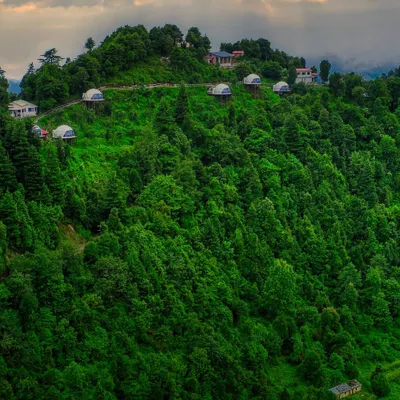 This is a drone photograph of the Keekoo Stays domes in Kanatal, nestled within an apple orchard on a hilltop. The image is composed to highlight the seclusion and the stunning view of the Himalayan range.