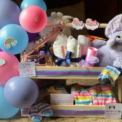 This two-level newborn essentials hamper is both beautiful and functional. The top tier holds diapers and a plush toy, while the bottom has neatly folded baby clothes and socks, all decorated with a "Promoted to Mommy" sticker.