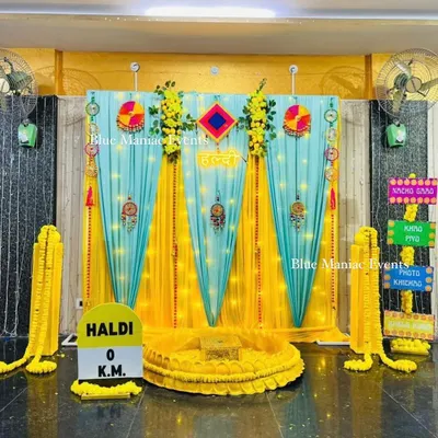 A complete Haldi ceremony setup designed for a home space. The backdrop combines bright yellow and turquoise drapes with traditional hangings and fun, quirky signs like 'Khao Piyo Photo Khichao'. The central seating area is defined by a low, decorated platform, ready for the ceremony.