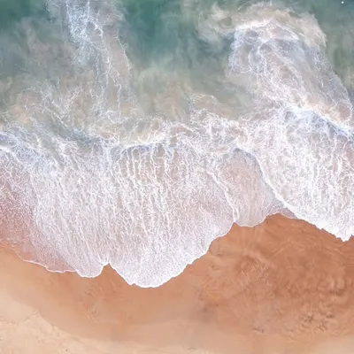 A top-down drone photograph of gentle waves washing over a sandy shore, creating a beautiful, abstract pattern.