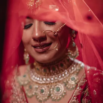 A classic, veiled bridal portrait. The bride's serene smile is captured through her delicate red dupatta, highlighting her stunning jewelry and the joy of the moment.