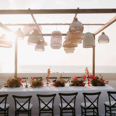 A stunning long-table setup for a beachside dinner in Goa. The scene is set against the ocean, with hanging rattan lamps and vibrant tropical floral runners creating a relaxed yet sophisticated atmosphere.