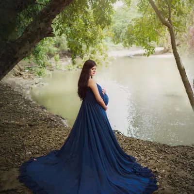 A serene moment captured by the water's edge. The long, flowing train of the blue maternity gown spreads out beautifully, creating a striking contrast with the natural surroundings.