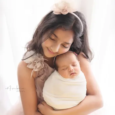 The beginning of a lifelong friendship. An older sister gently holds her swaddled newborn sibling, her expression full of love and tenderness.