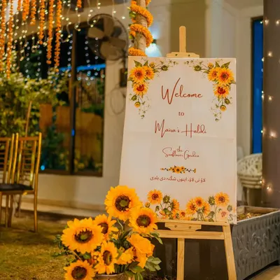 We make sure every detail of your celebration is perfect, day or night. This sunflower-themed welcome sign for an evening Haldi function is complemented by the warm glow of fairy lights and traditional marigold strings.