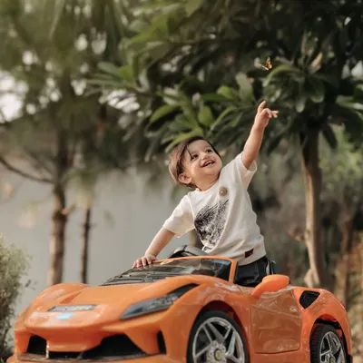 A little driver taking a break from his adventure. This outdoor shot with the orange sports car captures a moment of pure, joyful play against a beautiful green backdrop.