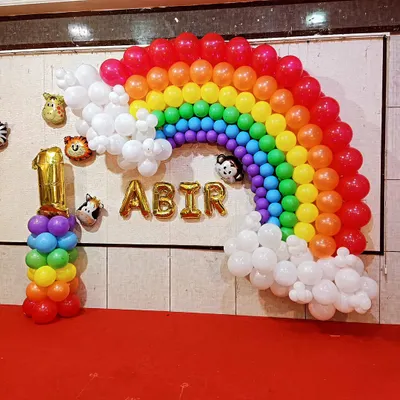 A wall-mounted rainbow balloon arch with fluffy white clouds, creating a beautiful photo backdrop. This setup for a first birthday includes animal head foil balloons peeking out of the clouds and the name "ABIR" in gold letters.