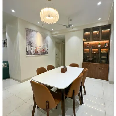 An elegant dining space featuring a white marble-top table, comfortable leatherette chairs, and a stunning feather chandelier. The glass crockery unit with internal lighting adds a touch of sophistication.
