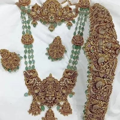 This is a magnificent antique bridal set featuring intricate carvings, goddess motifs, and green bead accents. It includes a choker, a long necklace, a statement vaddanam, and matching earrings.