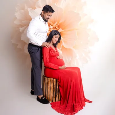 An intimate studio moment between expecting parents. The mom-to-be is seated while her partner stands by her side, set against a simple, elegant giant flower backdrop.
