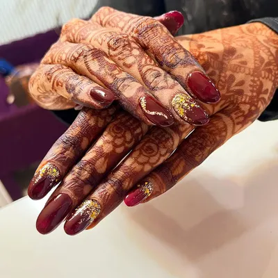 A stunning example of bridal nails designed to enhance the beauty of henna. This set features a rich maroon polish with gold glitter accents that sparkle against the intricate mehndi pattern.
