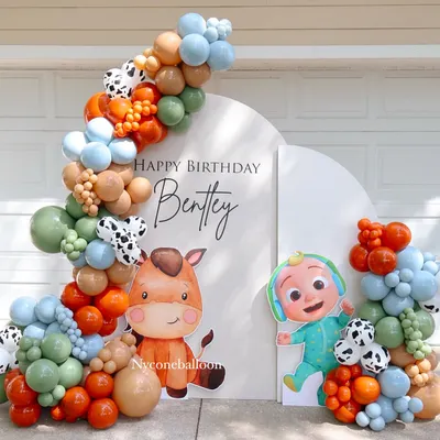 This modern Cocomelon setup for Bentley's birthday was arranged outdoors. It features a stylish arched backdrop and an organic balloon garland in a unique color scheme of sage green, orange, and tan with cow-print accents.