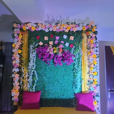 A close-up of a home Haldi backdrop, showing the combination of a green foliage panel with a border of colorful flowers and bright pink cushions.