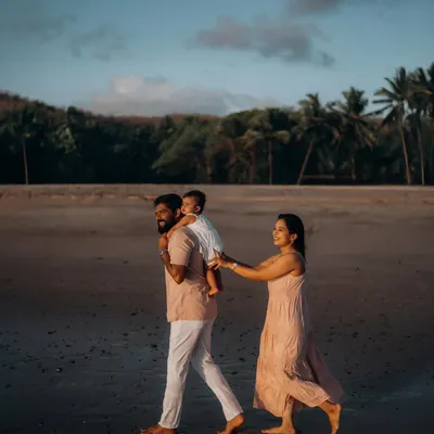 A father gives his child a shoulder ride as they walk along the wet sand at dusk, with the mother holding on playfully. This candid shot captures the simple, joyful moments of a family beach walk.