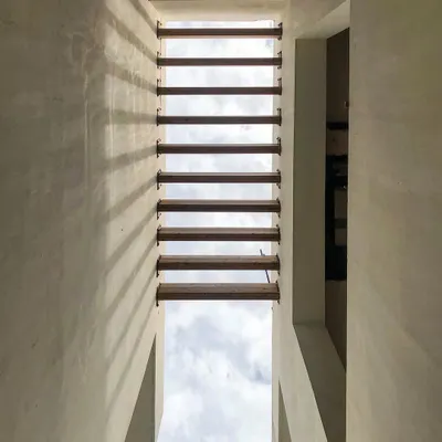 Looking up through a narrow void in a building, where wooden louvers create a rhythmic pattern against the sky. This detail is an example of how we use architectural elements to frame nature and play with light and shadow.