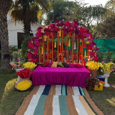 A beautiful outdoor mehndi setup with a vibrant floral arch in shades of pink and red. The decor includes traditional marigold tassels and a comfortable seating area on the lawn.