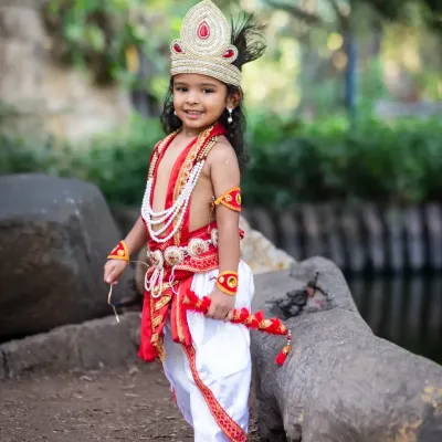 A full-length portrait of a young boy dressed as Lord Krishna, holding a flute and posing confidently in a park.