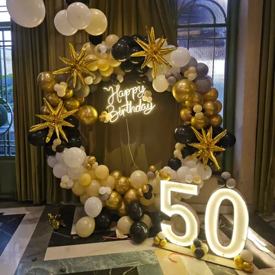 An elegant 50th birthday decoration featuring a black, gold, and white balloon ring, a 'Happy Birthday' neon sign, and large light-up '50' numbers.