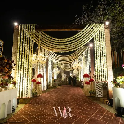 We designed this breathtaking entrance to blend rustic charm with traditional magic. A carpeted walkway is flanked by pillars draped in cascading white mogra, with chandeliers adding a regal glow, creating an enchanting prelude to the wedding celebration.