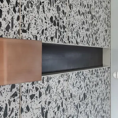 A close-up of the copper cuboid wedged into the terrazzo paneling in 'A Wedged Haus'. The reflective nature of the polished copper contrasts sharply with the matte, composite texture of the terrazzo stone.