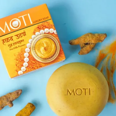 A clean product photograph for e-commerce, featuring a Moti luxury soap bar, its packaging, and the key ingredient of raw turmeric, all set against a solid blue background for maximum clarity.