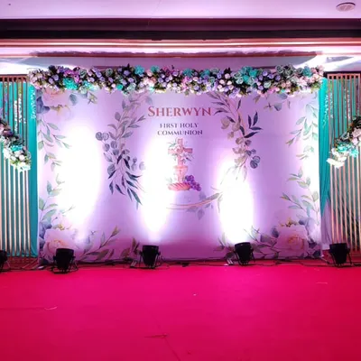An elegant and formal stage for a First Holy Communion celebration. The personalized backdrop, flanked by decorative panels and topped with a floral garland, creates a perfect focal point for photos and blessings.