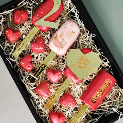 This anniversary gift box is designed for a truly personal celebration. It includes custom-engraved cakesicles and heart-shaped chocolates, all elegantly arranged on a bed of shredded paper.
