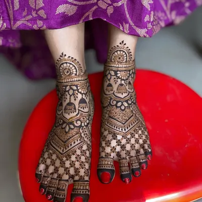 A full-coverage bridal foot mehandi design featuring a dense checkerboard pattern, floral details, and hanging jhumka motifs.