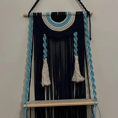 I love creating pieces that are both beautiful and useful. This evil eye themed wall shelf does just that, combining a protective symbol with a practical wooden shelf. The deep navy blue and sky blue cords with twisted hangers make it a unique piece.
