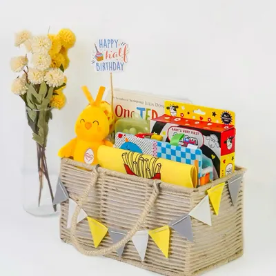 A 'Happy Half Birthday' themed baby hamper, beautifully arranged in a rustic basket. The bright yellow and grey color scheme is modern and cheerful.