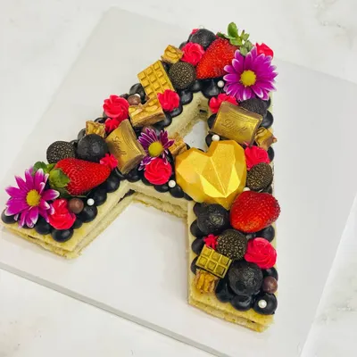 A stunning monogram cake in the shape of the letter 'A'. This one is loaded with fresh strawberries, blueberries, Lindt chocolates, and a geometric chocolate heart.