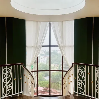 This interior view highlights a grand, double-height window that floods the space with natural light. The design is a fusion of classic and contemporary, with ornate white railings contrasting against dark walls and a circular skylight above.