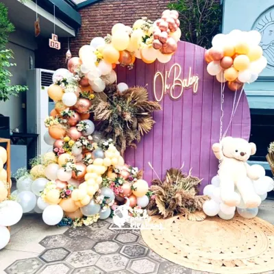 This rustic-themed baby shower featured a beautiful photo backdrop with a purple wooden panel, an 'Oh Baby' neon sign, and a large teddy bear floating on a cloud of balloons. The look was softened with pampas grass and a jute rug.