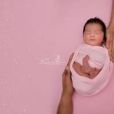 Held safely in her parents' hands. This classic newborn pose on a soft pink background highlights just how tiny and precious your baby is in their first few days.