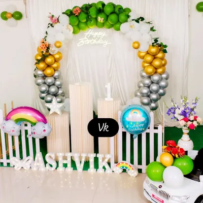 This first birthday setup features a circular balloon arch in green, gold, and silver. It includes a neon sign, marquee name lighting, and fun props like a rainbow balloon and a mini car.