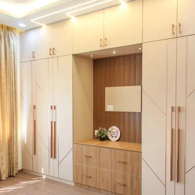 A full view of a spacious bedroom wardrobe in beige laminate with geometric routing and long, rose gold handles. The design includes a built-in dresser nook with a wood-paneled back and drawers.