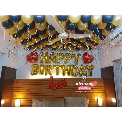 A classic bedroom birthday surprise. The ceiling is covered with black and gold balloons with dangling ribbons, complemented by gold foil "Happy Birthday" and heart-shaped balloons.