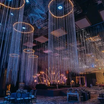 A wide view of the celestial-themed sangeet, where pillars of cascading crystals and illuminated rings hang from the ceiling. The design creates a magical, starry ambiance, with a neon lotus stage as the focal point for the evening's performances.