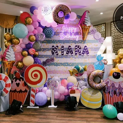A detailed look at a Candyland birthday theme, complete with a striped backdrop, neon "Happy Birthday" sign, and various props like giant cupcakes, lollipops, and donuts.