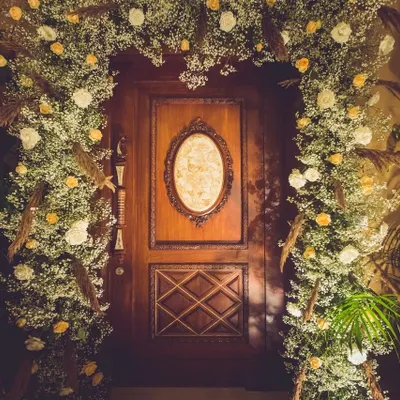 A grand doorway becomes a magical portal. We framed this beautiful wooden door with an arch of baby's breath, white roses, and dried pampas grass, creating an unforgettable entrance for a festive celebration.