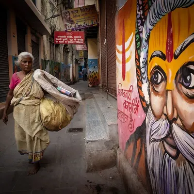 An elderly woman walks through a narrow lane in Varanasi, passing a vibrant mural of a sadhu. This image captures the intersection of daily life, art, and spirituality in the city's ancient streets.
