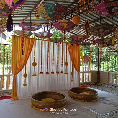 A full view of a terrace Haldi setup. The combination of colorful umbrellas, marigold hangings, and traditional 'urli' seating creates a perfect festive vibe.