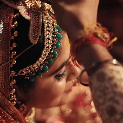 A close-up shot capturing the intricate details of an Indian bride's traditional headpiece, or 'matha patti'. The warm, golden light highlights the green beads and delicate craftsmanship as it is gently placed, symbolizing the transition from Miss to Mrs.