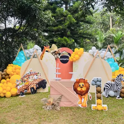 A full view of an outdoor safari camp theme for a first birthday. The setup includes teepees, a variety of animal cutouts like a lion and zebra, and balloon arrangements in earthy tones.