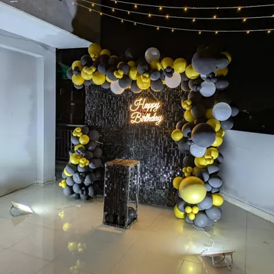 A full view of a black and yellow balloon arch framing a sparkling black sequin wall on a terrace. The setup includes a mirrored pedestal for the cake.