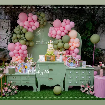A chic and modern first birthday setup with a sage green and pink color scheme. The decor features a stylish dessert table, organic balloon arches, and a neon '1' sign, creating a fresh and beautiful photo corner.