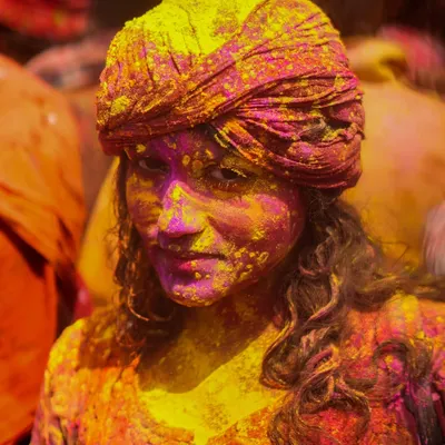 A stunning portrait of a woman at the Nandgaon Holi celebration, her face beautifully covered in yellow and pink gulal. Her direct gaze captures the joyful and immersive spirit of the festival.