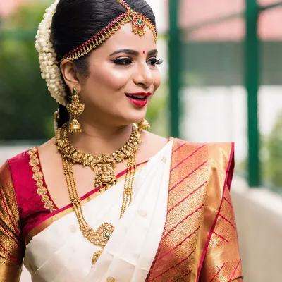 A classic South Indian bridal portrait. The look features sharp winged eyeliner, a bright red lip, and a neat hair bun adorned with fresh jasmine flowers, perfectly complementing her traditional silk saree and gold jewellery.