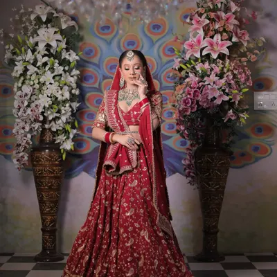 A full-length portrait of the bride in her magnificent red lehenga, standing between two large floral arrangements. This shot captures the grandeur of the entire bridal ensemble against a vibrant peacock-themed backdrop.
