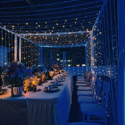 A magical rooftop dinner setup under a canopy of fairy lights. The blue ambient lighting and long table setting create a lavish and dreamy atmosphere for a special occasion.